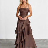 A woman modeling a brown strapless maxi dress with a ruched bodice and layered ruffle skirt, standing against a light gray studio wall.