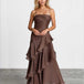 A woman modeling a brown strapless maxi dress with a ruched bodice and layered ruffle skirt, standing against a light gray studio wall.
