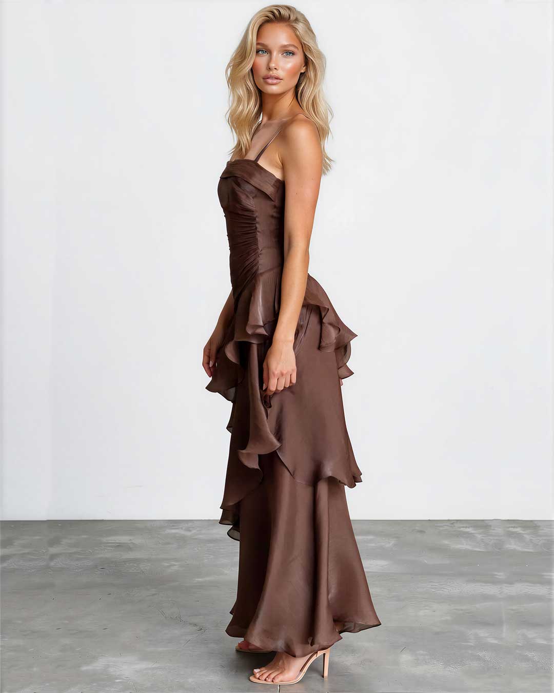 A blonde model wearing a brown ruffled halter-neck maxi dress with a structured bodice and layered skirt, standing against a white studio wall and gray floor.