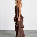 A blonde model wearing a brown ruffled halter-neck maxi dress with a structured bodice and layered skirt, standing against a white studio wall and gray floor.