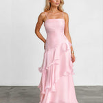 A woman modeling a light pink spaghetti-strap maxi dress with a ruched bodice and layered ruffle skirt, standing against a white studio wall and gray floor.