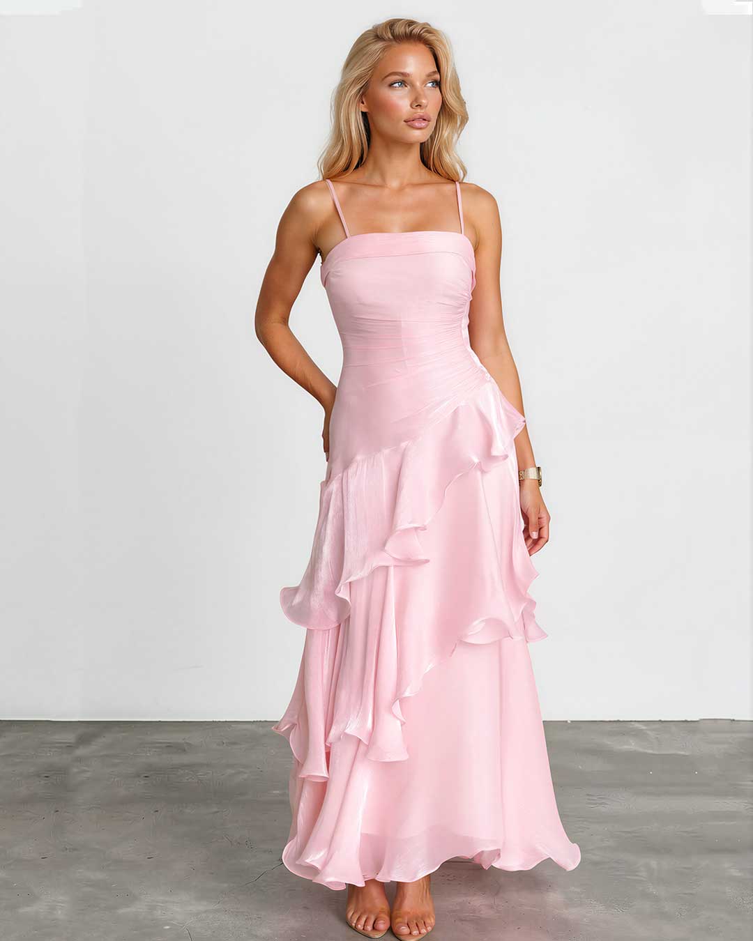 A woman modeling a light pink spaghetti-strap maxi dress with a ruched bodice and layered ruffle skirt, standing against a white studio wall and gray floor.