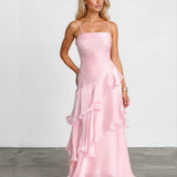 A woman modeling a light pink spaghetti-strap maxi dress with a ruched bodice and layered ruffle skirt, standing against a white studio wall and gray floor.