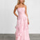 A woman modeling a light pink spaghetti-strap maxi dress with a ruched bodice and layered ruffle skirt, standing against a white studio wall and gray floor.