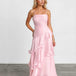 A woman modeling a light pink spaghetti-strap maxi dress with a ruched bodice and layered ruffle skirt, standing against a white studio wall and gray floor.