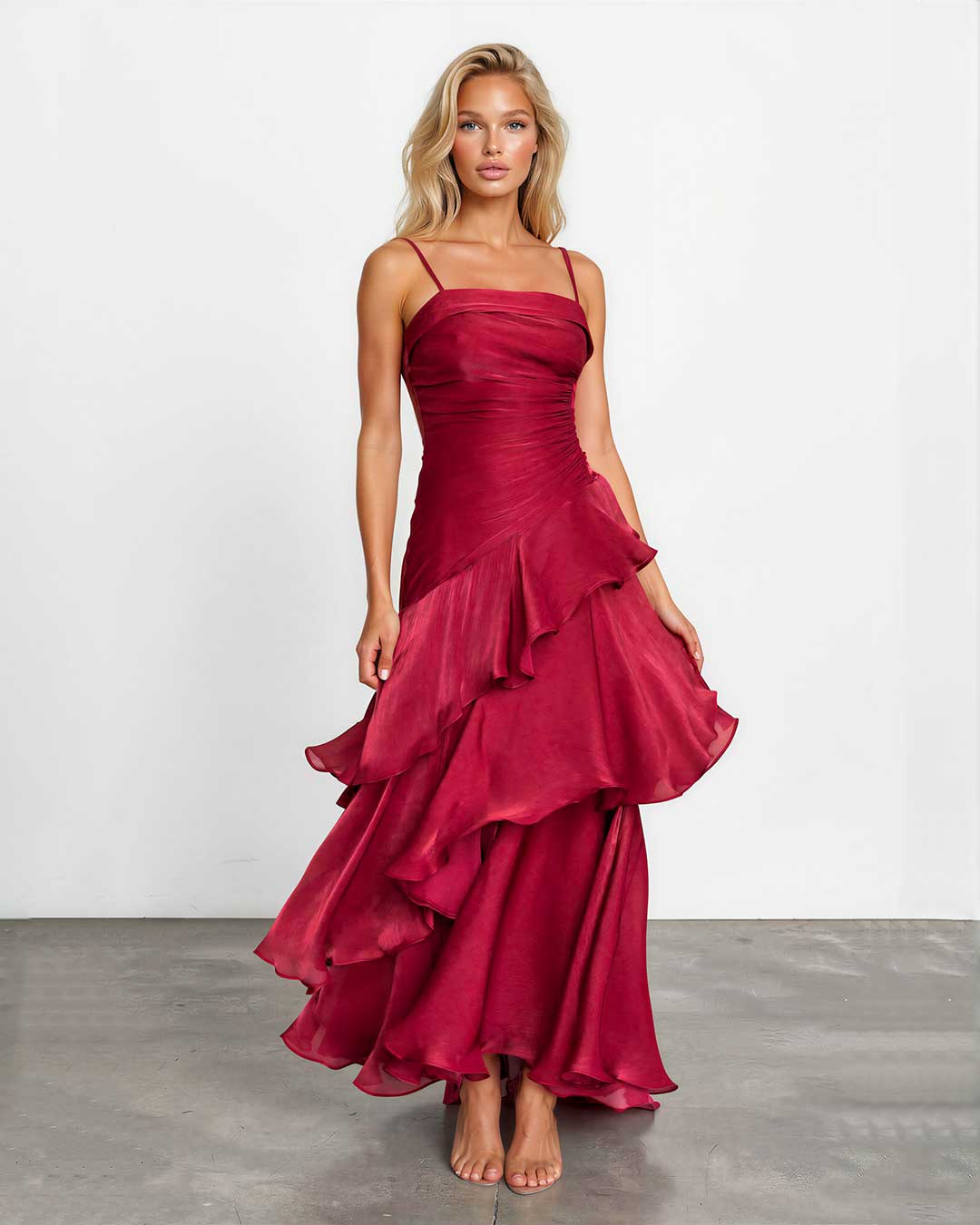 A woman modeling a sleeveless, tiered maxi dress in deep burgundy with a satin-like sheen, standing against a plain white studio wall on a gray floor.
