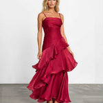 A woman modeling a sleeveless, tiered maxi dress in deep burgundy with a satin-like sheen, standing against a plain white studio wall on a gray floor.