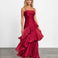 A woman modeling a sleeveless, tiered maxi dress in deep burgundy with a satin-like sheen, standing against a plain white studio wall on a gray floor.