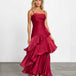 A woman modeling a sleeveless, tiered maxi dress in deep burgundy with a satin-like sheen, standing against a plain white studio wall on a gray floor.