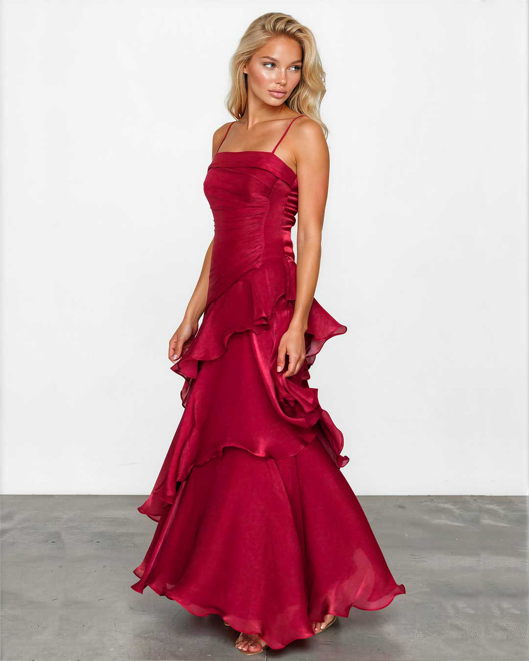 A woman modeling a floor-length burgundy satin gown with spaghetti straps, a ruched bodice, and dramatic layered ruffles, standing against a white studio wall.