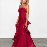 A woman modeling a floor-length burgundy satin gown with spaghetti straps, a ruched bodice, and dramatic layered ruffles, standing against a white studio wall.