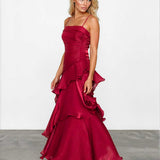 A woman modeling a floor-length burgundy satin gown with spaghetti straps, a ruched bodice, and dramatic layered ruffles, standing against a white studio wall.