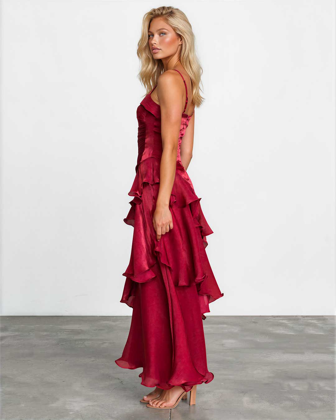 A blonde model wearing a deep red, multi-tiered ruffled maxi dress with spaghetti straps, standing in profile against a white studio wall and gray floor.