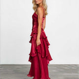 A blonde model wearing a deep red, multi-tiered ruffled maxi dress with spaghetti straps, standing in profile against a white studio wall and gray floor.