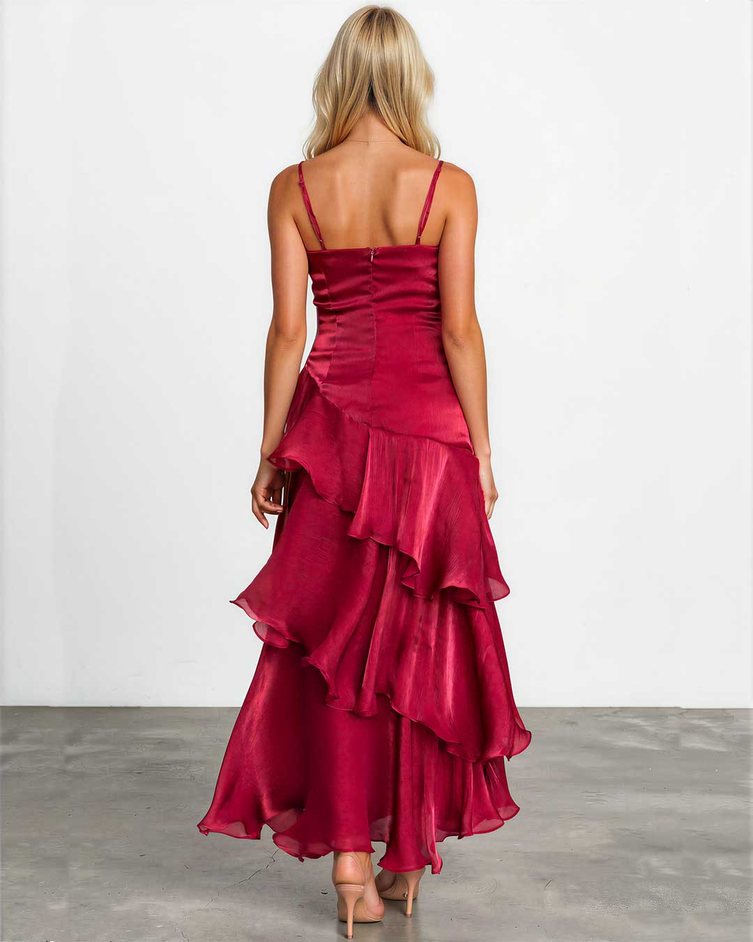 A woman viewed from behind wearing a sleeveless, deep burgundy maxi dress with thin straps, a structured ruched bodice, and a layered ruffled skirt. She is standing on a gray concrete floor against a white studio wall.