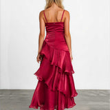 A woman viewed from behind wearing a sleeveless, deep burgundy maxi dress with thin straps, a structured ruched bodice, and a layered ruffled skirt. She is standing on a gray concrete floor against a white studio wall.