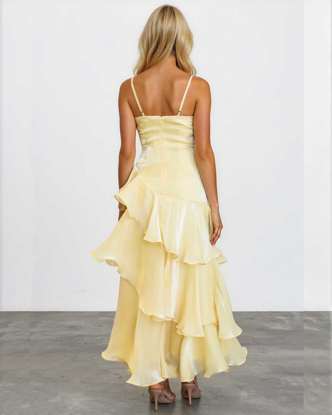A woman viewed from behind wearing a pale yellow ruffled spaghetti-strap dress with a structured bodice and layered skirt, standing against a plain light gray wall and concrete floor.