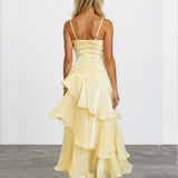 A woman viewed from behind wearing a pale yellow ruffled spaghetti-strap dress with a structured bodice and layered skirt, standing against a plain light gray wall and concrete floor.