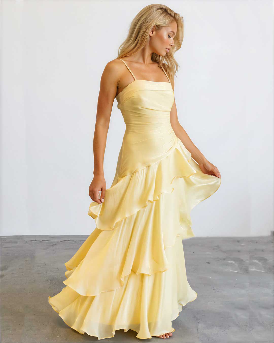 A woman modeling a pale yellow sleeveless maxi dress with thin straps, a fitted ruched bodice, and a layered ruffled skirt, standing against a plain light gray studio background.