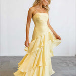 A woman modeling a pale yellow sleeveless maxi dress with thin straps, a fitted ruched bodice, and a layered ruffled skirt, standing against a plain light gray studio background.
