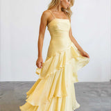 A woman modeling a pale yellow sleeveless maxi dress with thin straps, a fitted ruched bodice, and a layered ruffled skirt, standing against a plain light gray studio background.