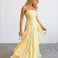 A woman modeling a pale yellow sleeveless maxi dress with thin straps, a fitted ruched bodice, and a layered ruffled skirt, standing against a plain light gray studio background.