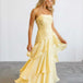 A woman modeling a pale yellow sleeveless maxi dress with thin straps, a fitted ruched bodice, and a layered ruffled skirt, standing against a plain light gray studio background.