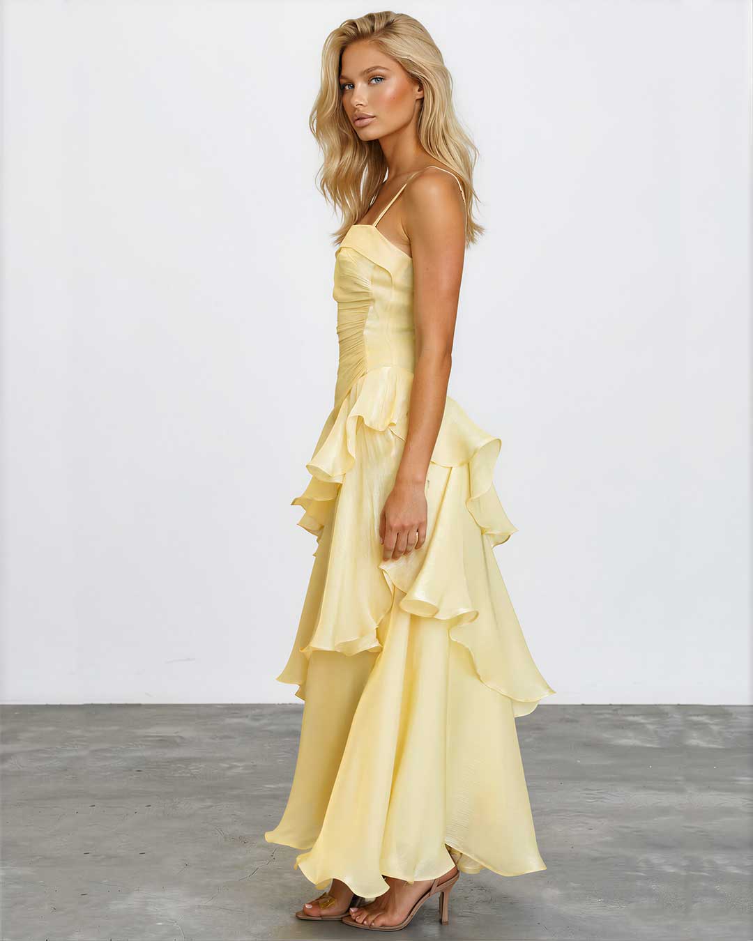 A blonde model wearing a pale yellow ruffled spaghetti-strap maxi dress with a tiered skirt, standing in profile against a white studio wall and gray floor.