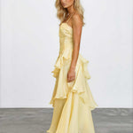 A blonde model wearing a pale yellow ruffled spaghetti-strap maxi dress with a tiered skirt, standing in profile against a white studio wall and gray floor.
