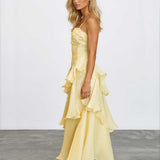 A blonde model wearing a pale yellow ruffled spaghetti-strap maxi dress with a tiered skirt, standing in profile against a white studio wall and gray floor.