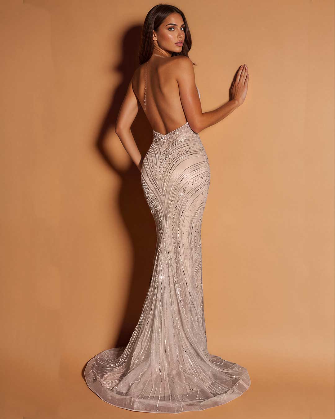 A woman modeling a backless, floor-length mermaid gown with intricate bead and sequin embellishments, posed against a warm tan studio backdrop.