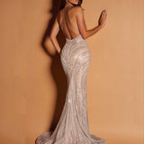 A woman modeling a backless, floor-length mermaid gown with intricate bead and sequin embellishments, posed against a warm tan studio backdrop.