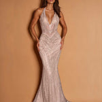 A woman modeling a floor-length, sleeveless halter gown with a plunging neckline and dense beaded embellishment, posed against a warm tan studio backdrop.