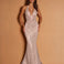 A woman modeling a floor-length, sleeveless halter gown with a plunging neckline and dense beaded embellishment, posed against a warm tan studio backdrop.