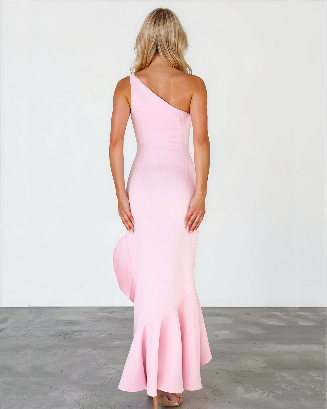 A woman stands with her back to the camera wearing a light pink one-shoulder mermaid-style gown with a flared ruffled hem, photographed against a plain light wall and gray floor.