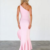 A woman stands with her back to the camera wearing a light pink one-shoulder mermaid-style gown with a flared ruffled hem, photographed against a plain light wall and gray floor.