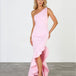 A blonde model wears a light pink one-shoulder midi dress with a ruffled asymmetrical hem, paired with clear high-heeled sandals, standing against a white studio wall and gray floor.