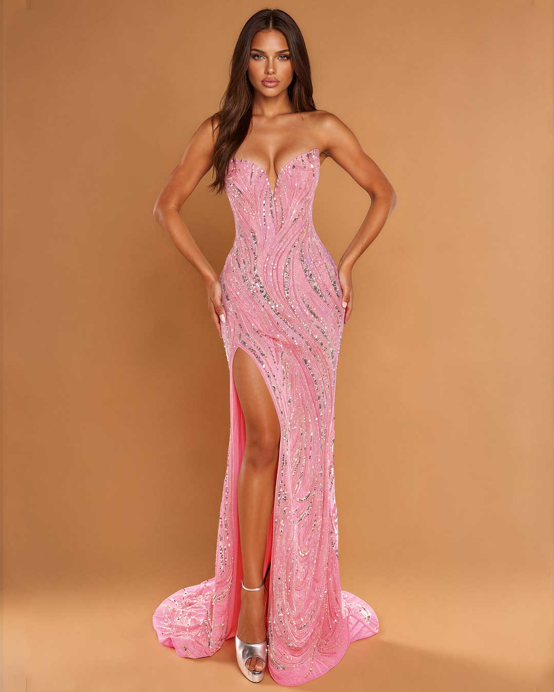 A woman models a strapless pink evening gown with silver sequin embellishments, a sweetheart neckline, and a high thigh slit, set against a solid tan studio background.