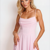 A blonde model wearing a light pink midi-length spaghetti-strap dress with a draped bodice and a flowing skirt, posed against a clean white studio background.