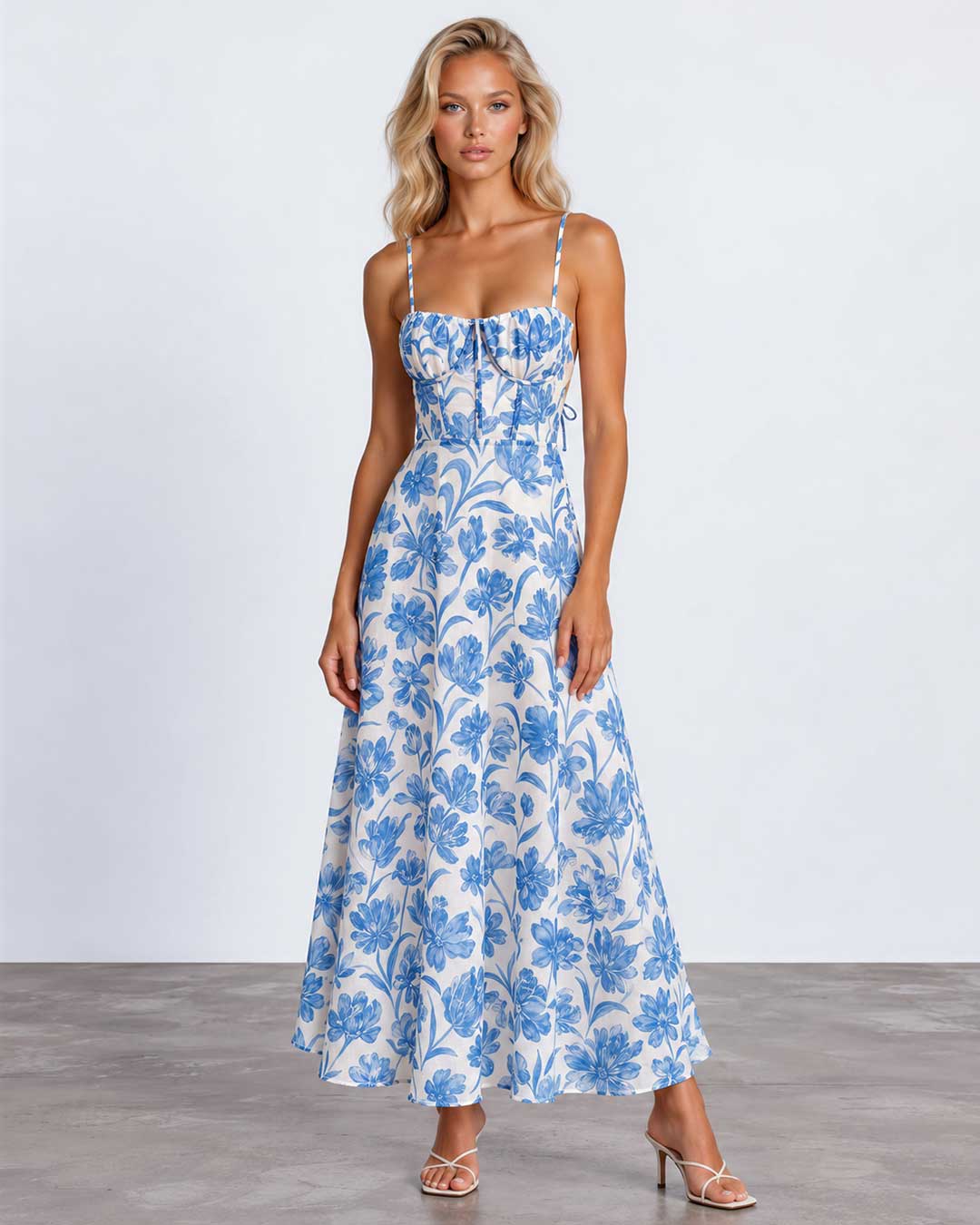 A woman modeling a sleeveless midi dress with thin straps, a gathered sweetheart neckline, and a blue floral toile pattern on a white background, standing against a light gray studio backdrop.
