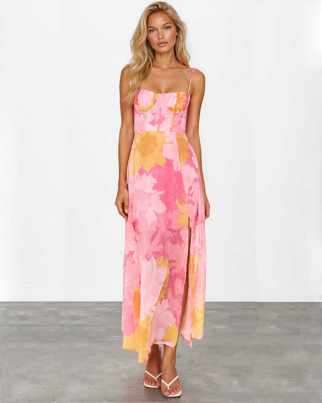 A woman modeling a sleeveless maxi dress with a pink and yellow floral print, standing against a white studio wall and gray floor.