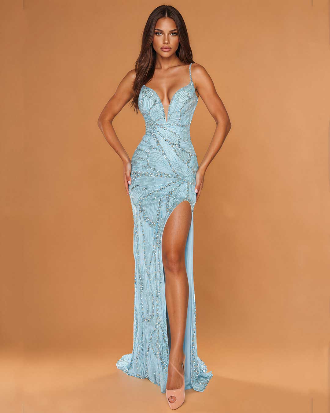 A woman modeling a floor-length light blue evening gown with thin straps, a deep V-neckline, a high thigh slit, and an embellished draped bodice, posed against a warm tan studio backdrop.