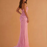 A woman modeling a floor-length pink gown with a structured lace-up back and a slight train, photographed against a warm tan studio backdrop.