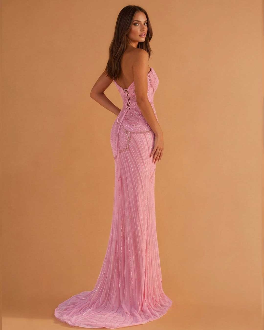 A woman modeling a floor-length pink gown with a structured lace-up back and a slight train, photographed against a warm tan studio backdrop.