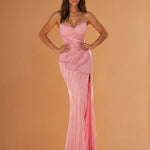 A woman modeling a floor-length pink gown with thin straps, a fitted ruched bodice, and a draped train against a warm peach studio background.