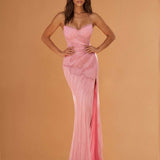 A woman modeling a floor-length pink gown with thin straps, a fitted ruched bodice, and a draped train against a warm peach studio background.