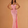 A woman modeling a floor-length pink gown with thin straps, a fitted ruched bodice, and a draped train against a warm peach studio background.