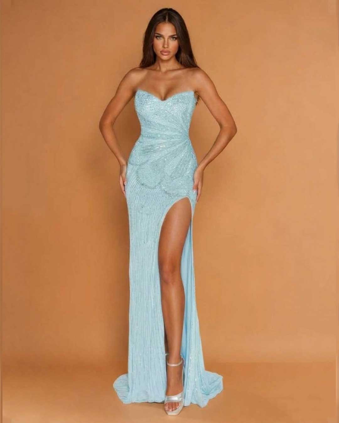A woman modeling a strapless light blue sequined gown with a sweetheart neckline, ruched bodice, high thigh slit, and floor-length train against a warm tan studio backdrop.