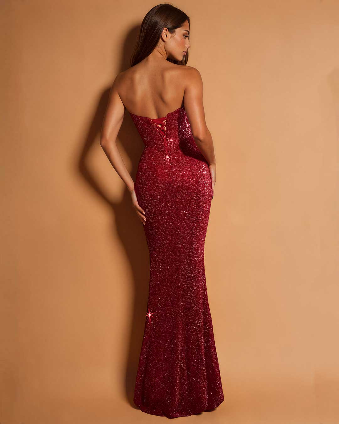 A woman modeling a strapless, deep V-neck, floor-length mermaid gown covered in sparkling burgundy sequins, shown from the back against a warm tan studio backdrop.