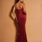 A woman modeling a strapless, deep V-neck, floor-length mermaid gown covered in sparkling burgundy sequins, shown from the back against a warm tan studio backdrop.